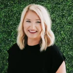 Smiling woman with blonde hair wearing a black top against a green leafy background.