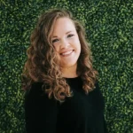 Smiling woman with curly hair in front of green foliage.