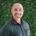 Smiling man in a black polo shirt with green foliage background.
