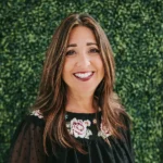 A smiling woman with long hair poses in front of a leafy green background.