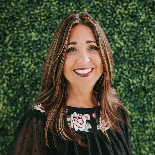 A smiling woman with long hair poses in front of a leafy green background.