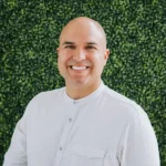 Smiling bald man in a white shirt against a leafy green background.