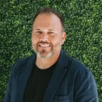 Smiling man in a dark blazer against a leafy green background.