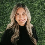 Smiling woman with long blonde hair in front of a greenery wall.