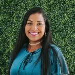 A woman with long dark hair smiles warmly against a leafy green background.