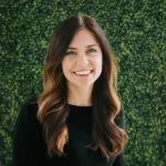 Smiling woman with long wavy hair in front of green foliage.