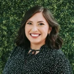 Smiling woman with dark hair standing in front of a leafy green background.