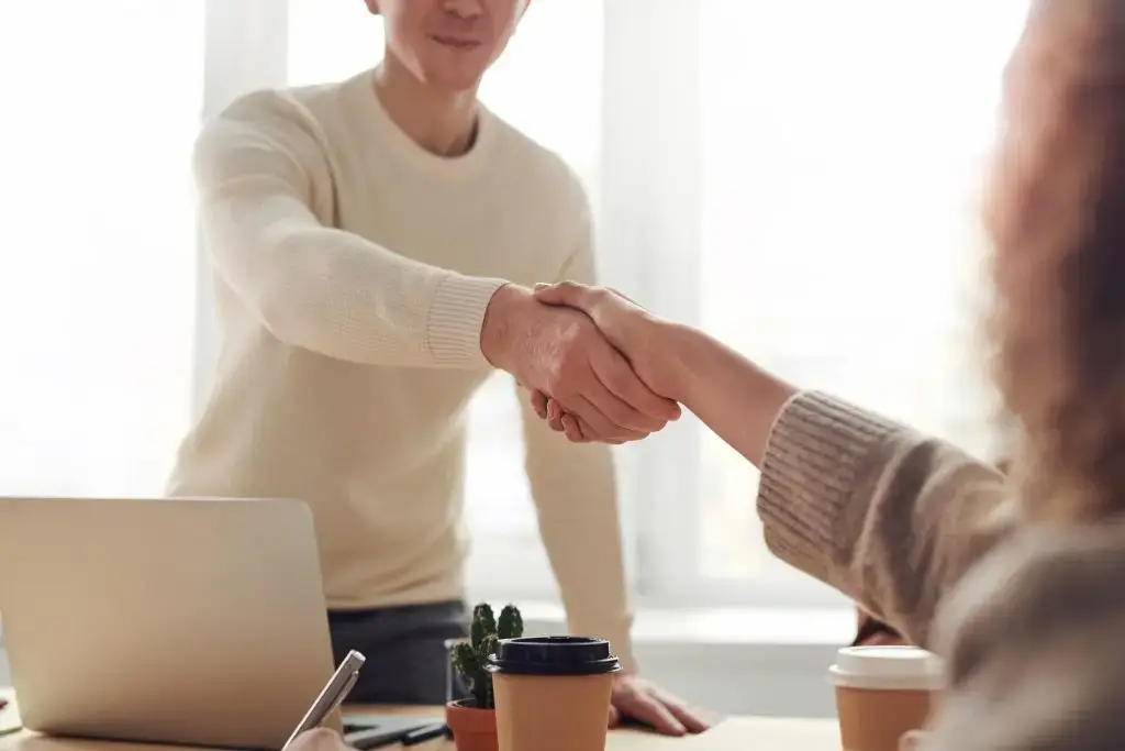 Two people shaking hands over a desk in a bright office.
