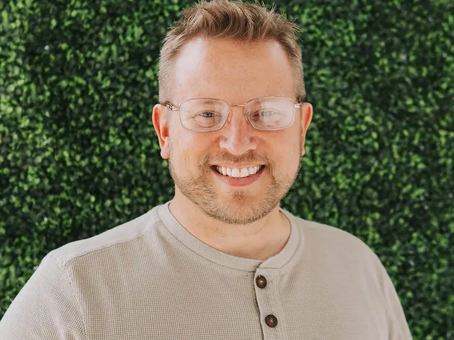 Smiling man with glasses in front of green leafy background.
