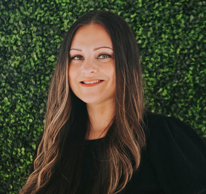 Sarah McCormick is smiling, wearing a black shirt, and standing in front of a green background.