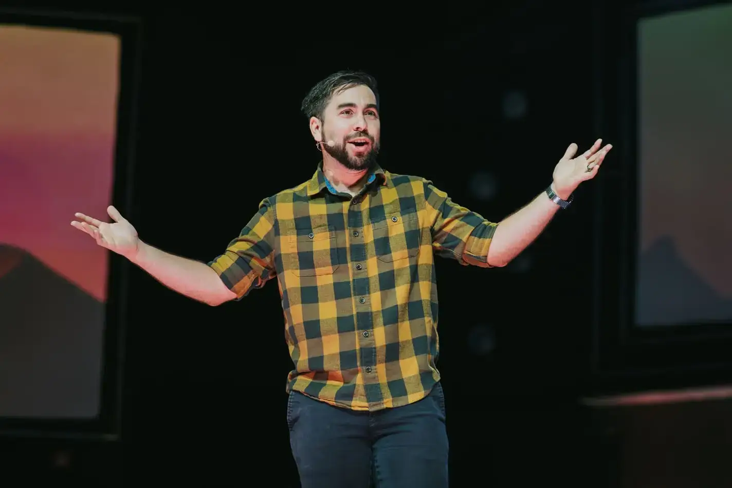 Man in a yellow plaid shirt speaking animatedly with open arms on stage.