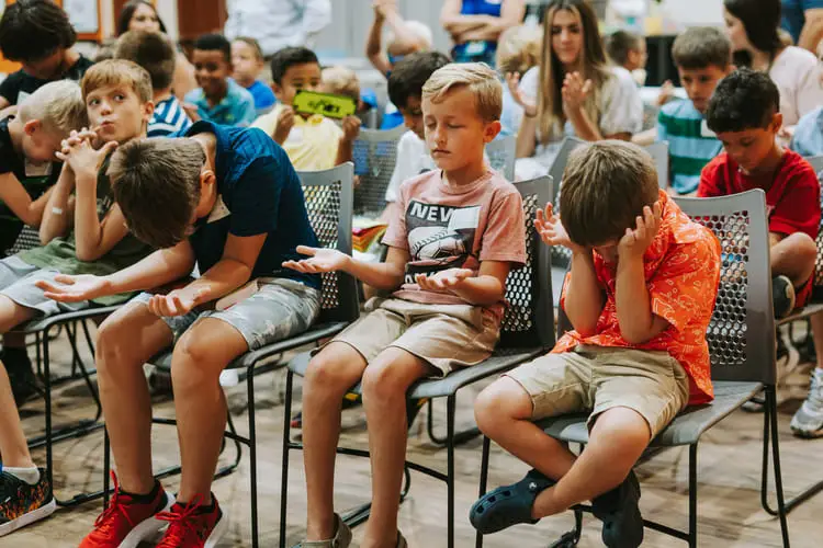Children sitting in chairs, some looking bored or upset, others distracted.