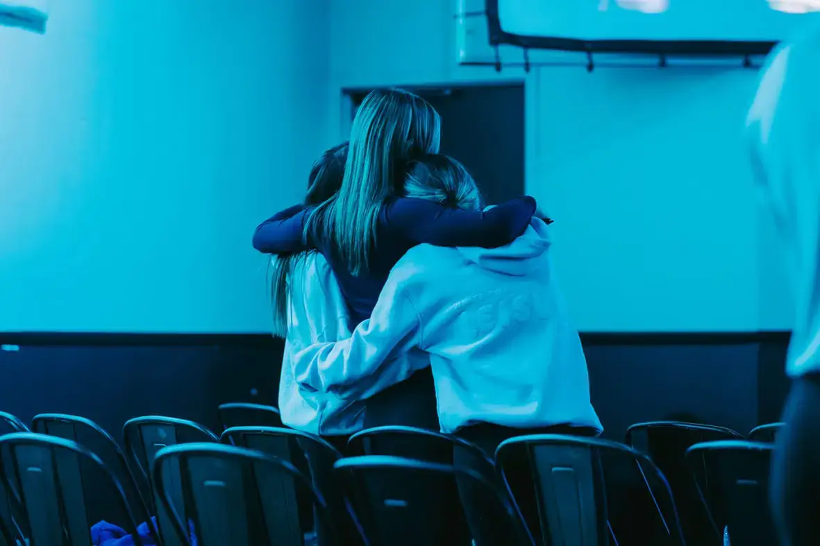 Two people embracing in a dimly lit room with chairs.